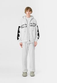 Sudadera blanca con cremallera y logo negro en las mangas, combinada con pantalones de chándal blancos y calzado texturizado de color gris claro. El modelo está de pie contra un fondo blanco.