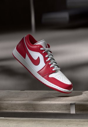 Rood-witte Nike Air Jordan 1 Low sneaker met grijze veters, schuin in de lucht boven een betonnen ondergrond in zacht licht.