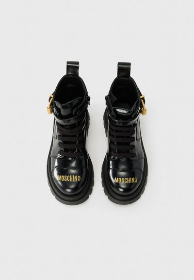 Stivaletti alla caviglia in pelle verniciata nera con logo dorato "MOSCHINO", lacci neri, zip laterale e suole in gomma nera spessa. Presentano un anello a forma di tab.