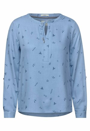 Blouse bleu clair à manches longues avec petits gribouillis bleu marine, encolure en trou de serrure et détail noué, avec des mots comme "amour" et des papillons.
