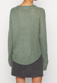 Vihreä neulepaita, jossa on tekstuuri, pitkät hihat ja pyöreä helma. Paitaa on käytetty harmaan farkkuhameen päällä, jossa on ribbaatut hihansuut.