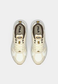 Zapatillas Fila de color crema, con cuero texturizado, rayas amarillas y negras, sistema de cordones en la parte frontal y un interior acolchado marrón, que presenta un logotipo destacado.