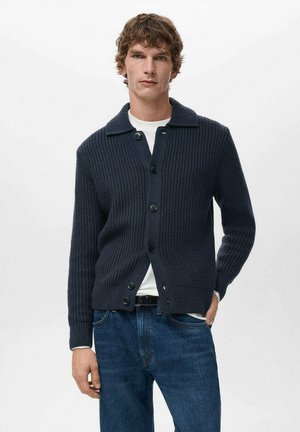 Cardigan azul marino acanalado con cuello con solapa, cinco botones negros y un ajuste relajado, combinado con vaqueros azules y una camisa blanca debajo.