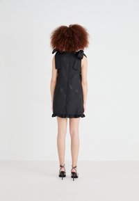 Black sleeveless dress with ruffled hem, floral texture, and bow accents on shoulders. Paired with strappy black high heels.