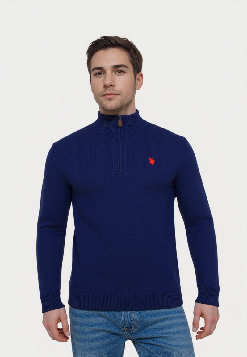 Marineblauer Strickpullover mit einem Reißverschlusskragen und gerippten Bündchen, versehen mit einem kleinen roten Logo auf der linken Brust, kombiniert mit hellblauen Jeans.