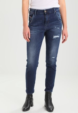 Femme portant un jean slim bleu foncé avec des empiècements usés, un haut blanc et des bottines noires à talons, debout devant un fond uni.