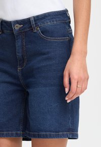 Tamsiai mėlynos spalvos džinsiniai šortai su lygiu paviršiumi, tradicinėmis kišenėmis ir auksinio siūlės siuvimu. Rankos laikomas mažas žiedas yra šalia.