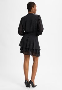 Schwarzes, gestuftes Kleid aus transparentem Stoff, mit langen Ärmeln und gerafftem Bund, ergänzt durch eine schmale Passform und hohe Absätze.