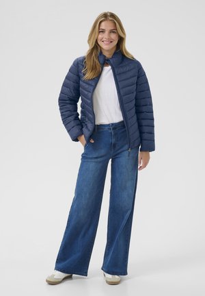 Glunderende vrouw met lang blond haar, gekleed in een blauwe gewatteerde jas, een wit shirt, brede spijkerbroeken en witte sneakers, met haar handen in de zakken.