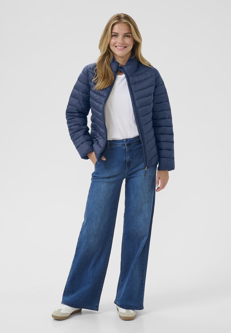 Glunderende vrouw met lang blond haar, gekleed in een blauwe gewatteerde jas, een wit shirt, brede spijkerbroeken en witte sneakers, met haar handen in de zakken.