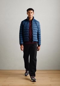 Blauwe pufferjack met horizontale stiksels, bordeauxrode gebreide trui eronder en zwarte broek. Draagt een paar sneakers met een patroon en poseert binnen.