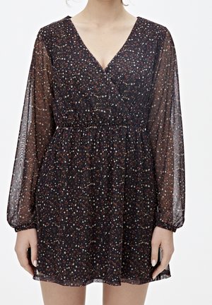 Vestido mini negro translúcido de manga larga con pequeño estampado floral multicolor y cintura elástica, llevado por una persona de pie, brazos relajados a los lados.
