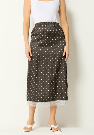 Vrouw draagt een zwarte midi-rok met witte polkadots en kanten rand, wit tanktopje en witte sandalen met hak, staand tegen een effen achtergrond.