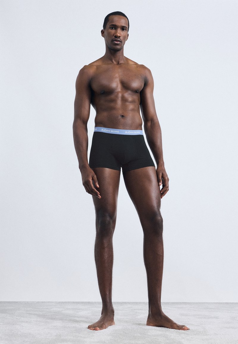 Μαύρα ανδρικά μποξεράκια με ανοιχτό μπλε ζώνη μέσης και λογότυπο. Το μοντέ λο έχει μυώδη σωματοδομή και στέκεται ξυπόλυτο μπροστά σε απλό φόντο.