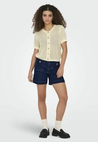 Top de punto color amarillo claro de manga corta con cierre de botones y borde festoneado, combinado con shorts de mezclilla oscuros que presentan bolsillos frontales y detalles dorados.