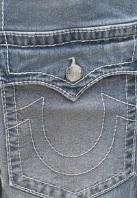 Dżinsy o jasnoniebieskim kolorze, z kontrastującymi białymi szwami i guzikiem z logo na tylnej kieszeni, podkreślone zakrzywionymi wzorami.