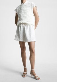 Top senza maniche in crochet bianco sovrapposto a una gonna corta bianca con motivo. Abbinato a sandali con tacco di colore chiaro. Tessuti morbidi e vestibilità casual.