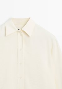 Camicia color crema chiaro con bottoni, realizzata in tessuto morbido, caratterizzata da un colletto tradizionale e sei bottoni sul davanti, con una texture liscia.