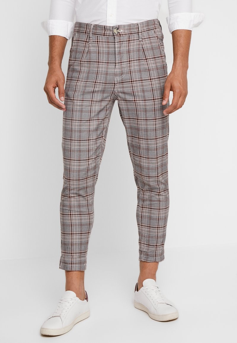 Homme portant un pantalon gris à carreaux ajusté avec jambes raccourcies, associé à des baskets blanches et une chemise blanche à boutons.