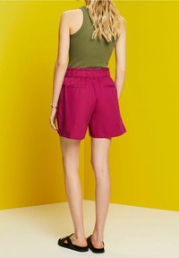 Shorts magenta con cintura elástica y dos bolsillos traseros, combinados con una camiseta sin mangas verde oliva, contra un fondo amarillo.