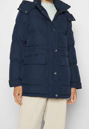 Femme portant une veste matelassée bleu marine avec poches à rabat, boutons-pression et capuche amovible, associée à un pantalon en velours côtelé crème.