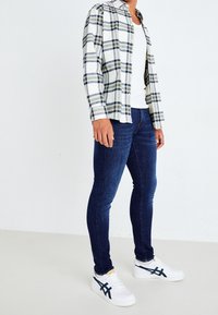 Rutigt flanellskjorta i vitt, marinblått och grönt; åtsittande mörkblå jeans; vita sneakers med marinblå detaljer. Avslappnad outfit, inga synliga accessoarer.