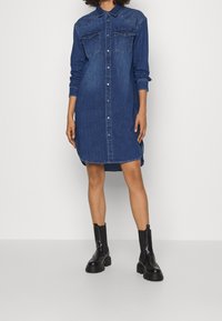 Denimskjortklänning i mörkblått med långa ärmar, knapplist, två bröstfickor, sidofickor och en avslappnad passform, kombinerad med svarta ankelskor.