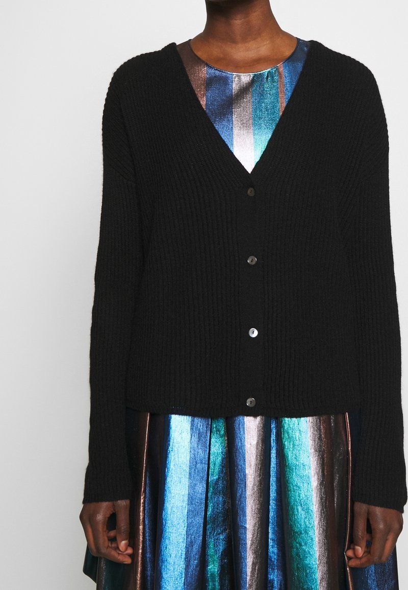 Černý žebrovaný kardigan s výstřihem do V, který má pět předních knoflíků. Nositý přes vícebarevné metalické pruhované šaty v modrých a bronzových tónech.