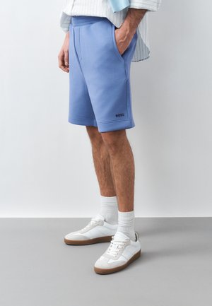 Férfi világoskék BOSS logós rövidnadrágban, fehér tornacipővel és fehér zokniban, egyszerű fehér és szürke háttér előtt állva.