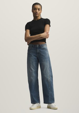 Junge Frau mit geflochtenem Haar steht mit verschränkten Armen da, trägt ein schwarzes, kurzes T-Shirt, weit geschnittene blaue Jeans und weiße Turnschuhe.