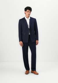 Sacou bleumarin închis, cu două rânduri de nasturi, cu o textură fină, asortat cu o cămașă albă și pantofi din piele maro, având o croială elegantă și ajustată.