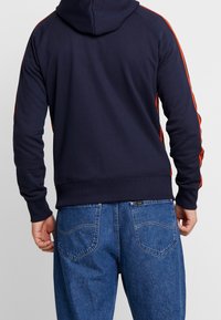 Persona che indossa un felpa navy con strisce rosse sulle maniche e jeans di jeans blu, mostrato da dietro su uno sfondo neutro.
