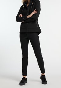 Svart zip-up jacka med huva, prydd med dekorativa pärlor och kontrasterande vit sömnad. Kombinerad med svarta skinny jeans och svarta sneakers.