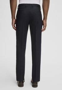 Dunkle Anzughosen aus strukturiertem Stoff, mit geradem Bein, Gürtelschlaufen und zwei Gesäßtaschen. Getragen mit braunen Schuhen.