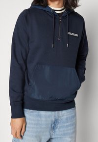 Felpa navy in morbido tessuto con tasca frontale in nylon, polsini a costine e zip a un quarto. Caratterizzata da una toppa con logo sul petto.