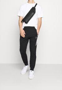 Černé tepláky se bočními pruhy, spárované s bílým tričkem. Model má na sobě texturovanou černou tašku přes rameno a bílé tenisky.