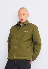 Veste en coton vert olive avec un col, des boutons-pressions et des coutures blanches ; elle dispose de deux poches latérales et d'un logo Timberland.