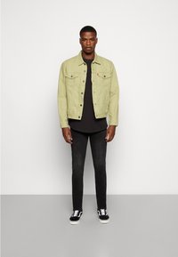 Hellgrüne Jeansjacke mit zwei Brusttaschen und Knopfverschluss, getragen über einem schwarzen Hemd, kombiniert mit schwarzen Jeans und schwarzen Vans Sneakern.