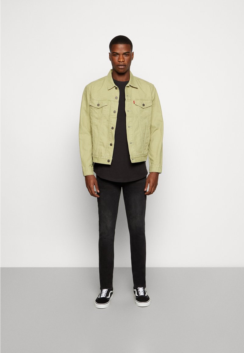 Hellgrüne Jeansjacke mit zwei Brusttaschen und Knopfverschluss, getragen über einem schwarzen Hemd, kombiniert mit schwarzen Jeans und schwarzen Vans Sneakern.