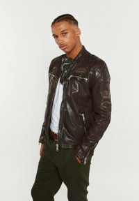 Veste de moto en cuir marron avec des accents matelassés aux épaules, des poches zippées et des écussons. Portée sur une chemise blanche et un pantalon vert.