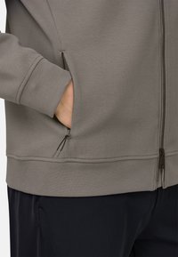 Main dans la poche latérale d'une veste taupe zippée avec tirette de fermeture éclair visible, portée avec un pantalon noir.