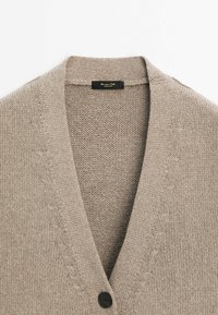 Cardigan beige en tricot avec un profond col en V et un seul bouton noir visible près du bas de l'encolure.