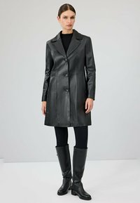 Czarny skórzany płaszcz z ostrym kołnierzem, zapięciem na guziki i pionowymi szwami. Noszony z czarnymi leginsami i wysokimi botkami. Gładka tekstura.