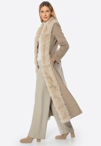 Beige jas met een faux fur kraag, lange mouwen en zakken. Gecombineerd met lichte broeken en chunky haklaarzen, waarmee zachte texturen worden getoond.