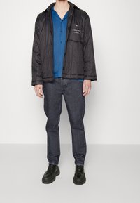 Chaqueta aislante negra con cremallera, acabado texturizado, camisa azul debajo, jeans de mezclilla oscura y zapatos negros de cordones con suelas gruesas.