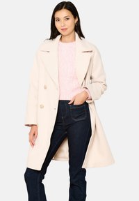 Kremowy wełniany płaszcz o podwójnym zapięciu, dużych klapach i dwóch przednich kieszeniach, noszony na różowym swetrze i ciemnych dżinsach.