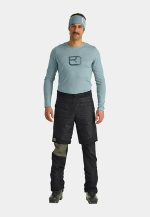 Helles blaues Langarmhemd mit schwarzem Logo, kombiniert mit schwarzen gesteppeten Shorts und schwarzen Hosen mit grünen Stoffakzenten.