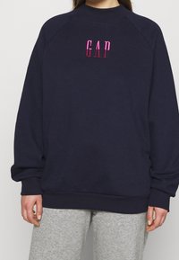 Laivastonsininen raglan-hihoilla varustettu collegepaita, jossa on vaaleanpunainen kirjailtu "GAP"-logo rinnassa. Pinta on teksturoitu, rento istuvuus ja resorireunaiset hihansuut.