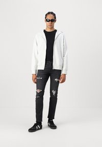 Svijetlosiva zip-up dukserica s prednjim džepom, crna majica s okruglim izrezom, iznjedrene crne traperice i crne tenisice s bijelim prugama.
