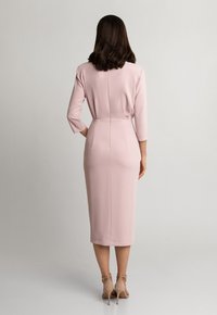 Dopasowana, sukienka do kolan w jasnoróżowym materiale z rękawami o długości trzech kwadransów i ukrytym zamkiem z tyłu, zestawiona z beżowymi szpilkami.